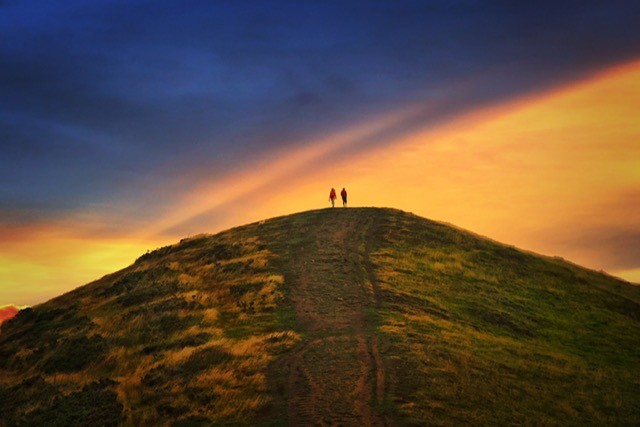 Photo of one of the Malvern hills with two walkers on top enjoying a dramatic orange and blue sky.