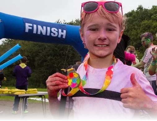 a young person prodly showing off a colourful medal, in front of a finish line.