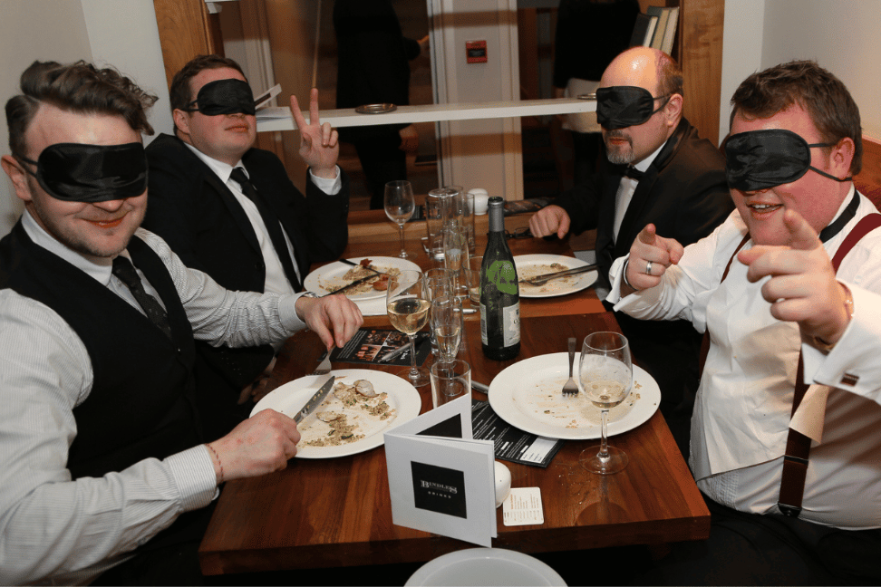 4 diners sat eating a meal with blindfolds on.